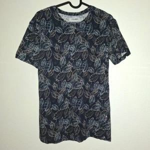 Blå t-shirt med bladmönster Strömberg - Snygg blå t-shirt från Strömberg i ekologisk bomull. T-shirten har ett all-over bladmönster i grått och beige, rund hals och korta ärmar. Perfekt för dig som gillar naturinspirerade prints och vill ha en chill och bekväm stil.