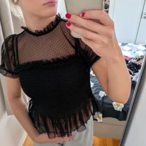 Svart meshblus med volang och prickar - Supersöt svart blus i mesh med små prickar och volangdetaljer vid hals, ärmslut och nederkant. Transparent upptill och på ärmarna, smockad över bysten och midjan för en tight passform. Perfekt för dig som gillar romantiska och trendiga detaljer.