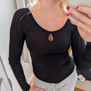 Svart ribbad långärmad topp - Svart ribbad topp med lång ärm och snyggt hål vid urringningen. Ärmarna har dekorativa hål längs hela längden som ger en cool detalj. Sitter tight och har en feminin passform, perfekt till jeans eller kjol.