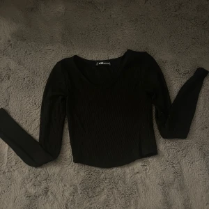 Svart ribbad långärmad topp FB Sister - En svart, ribbad långärmad topp från FB Sister i storlek XXS. Toppen har v-ringning och är croppad, vilket ger en snygg och modern look. Perfekt att matcha med höga jeans eller kjol för en stilren outfit.
