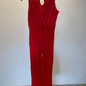 Röd byxdress från DKS, storlek S - Snygg röd byxdress från DKS med omlott framtill och ärmlös design. Byxdressen har resår i midjan för en skön passform och vida ben. Perfekt för dig som vill sticka ut med en färgstark och trendig look. Liten defekt vid axeln men går att sy utan problem. 