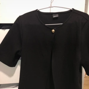 Svart kofta med guldig knapp Gina Tricot - Stilren svart kofta från Gina Tricot med kort ärm och rund halsringning. Koftan har en snygg guldig knapp framtill som detalj och är gjord i ett mjukt, stickat material. Perfekt att slänga över en topp för en clean look.