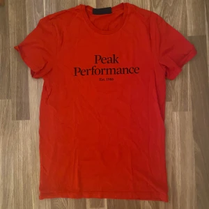 Röd Peak Performance t-shirt - Snygg röd t-shirt från Peak Performance med svart tryck på bröstet. Klassisk rund hals och korta ärmar, perfekt för en chill och sportig stil. Materialet känns mjukt och skönt mot huden. Enkelt och stilrent plagg som funkar året runt.