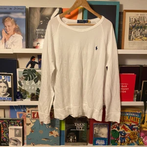 Vit långärmad tunn tröja Polo Ralph Lauren - Vit långärmad tröja från Polo Ralph Lauren med klassisk rund halsringning och broderad marinblå logga på bröstet. Tillverkad i mjuk bomull, perfekt för en clean och stilren look. Tröjan är enkel att matcha till det mesta.