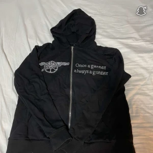 Svart Arsenal hoodie med strass - Svart hoodie med huva och dragkedja från Arsenal. Framsidan har texten 'Once a gunner always a gunner' och klubbens kanon-logga i strass. Perfekt för dig som älskar fotboll och vill visa ditt stöd för laget på ett stilrent sätt.