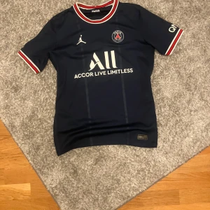 PSG Mbappé matchtröja Jordan L - Snygg PSG fotbollströja med Mbappé #7 på ryggen, officiell Jordan edition. Marinblå med vita tryck, röda och vita detaljer på krage och ärmslut. PSG-emblem på bröstet och Accor Live Limitless sponsortryck. Tillverkad i lätt polyester, perfekt för match eller träning. Storlek L i barn som motsvarar XS