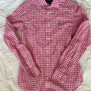 Rutig rosa skjorta från Ralph Lauren - Snygg rutig skjorta från Ralph Lauren Sport i rosa och vitt med klassisk krage och knappar framtill. Broderad blå logga på bröstet. Skjortan är långärmad och tillverkad i mjuk bomull, perfekt för en chill och stilren look.