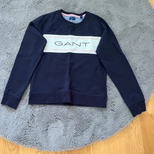 Mörkblå sweatshirt från GANT - Säljer en snygg mörkblå sweatshirt från GANT med vit panel och rosa detaljer över bröstet där det står GANT. Tröjan har rund halsringning, långa ärmar och är gjord i mjuk bomull. Perfekt för en chill och stilren look.