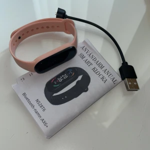 Rosa smartklocka AX6+ - Smartklocka från AX6+ med rosa silikonarmband och svart rektangulär display. Klockan har touchskärm och kommer med laddkabel och användarmanual. Perfekt för dig som vill ha koll på steg, puls och notiser.
