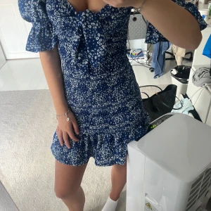 Blommig blå kortklänning Gina Tricot - Supersöt blå kortklänning från Gina Tricot med vita blommor, puffiga trekvartsärmar och volangkant nedtill. Tajt men luftig passform med en rosett framtill. Perfekt för festliga tillfällen 