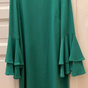 Grön klänning med volangärm Indiska - Supersnygg grön klänning från Indiska med långa ärmar och dubbla volanger vid ärmsluten. Rak passform och enkel design utan mönster, perfekt för dig som gillar statement-ärmar. Materialet känns mjukt och har lite struktur.