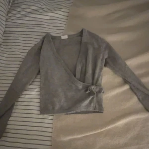 Grå omlottopp med knyt - Snygg grå omlottopp med lång ärm och knyt i sidan. Djup v-ringning och croppad passform gör den perfekt till både jeans och kjol. Mjuk och skön, passar dig som gillar stilrena plagg med en twist.