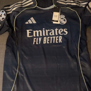 Real Madrid Mbappé #10 fotbollströja - Snygg marinblå Real Madrid fotbollströja från Adidas med Mbappé och nummer 10 på ryggen. Tröjan har korta ärmar, vita detaljer och klubbmärke på bröstet. Materialet är lätt och andas, perfekt för match eller träning. Champions League-märke på ärmen och klassisk Adidas-design. En äkta tröja direkt från adidas hemsida 