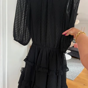 Svart kort volangklänning med prickar - Supersöt svart kortklänning med transparenta, prickiga ärmar och volanger i kjolen. Klänningen har öppen rygg och trekvartsärmar med resår. Tunt, lätt material som ger en luftig känsla och snyggt fall. 