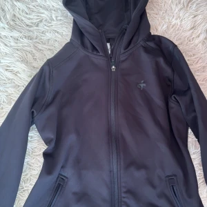 Svart vindjacka från Cross Sportswear - Svart vindjacka från Cross Sportswear med huva och hel dragkedja framtill. Jackan har två fickor med dragkedja och är fodrad med mjuk fleece på insidan. Perfekt för blåsiga dagar och enkel att matcha med allt.