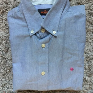 Ljusblå skjorta från Morris - Klassisk ljusblå skjorta från Morris med button down-krage och vita knappar. Broderad rosa logga på bröstet ger en snygg detalj. Skjortan är i bomull och har lång ärm, perfekt till jeans eller chinos.