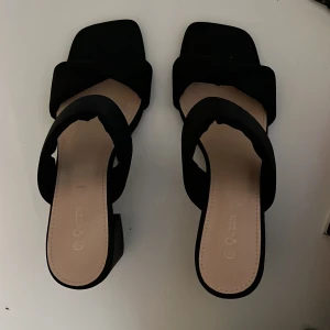 Svarta sandaler med blockklack - Stilrena svarta sandaler från Queen med breda remmar och fyrkantig tå. Skorna har en chunky blockklack som ger både höjd och stabilitet. Perfekta för dig som gillar minimalistisk och trendig design. Ovandelen är i mjukt syntetmaterial.