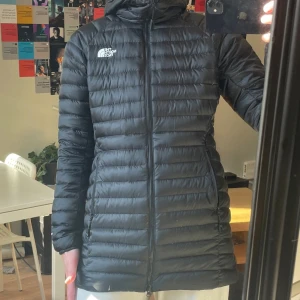 Svart dunjacka från The North Face I stl M oanvänt  - Säljer en svart, lång dunjacka från The North Face med huva och dragkedja framtill. Jackan är quiltad med horisontella sömmar och har en clean, sportig look. Perfekt för kalla dagar när du vill hålla värmen och ändå se snygg ut.