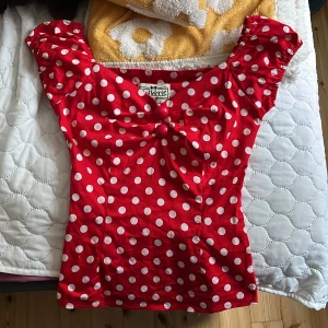 Röd topp  - Supersöt röd topp med vita prickar från Collectif. Modellen har korta puffärmar, två dekorativa knappar framtill och en söt rosettliknande detalj vid bysten. Toppen har en figurnära passform och dragkedja i sidan. Perfekt för dig som gillar retrostil! Säljer då den inte kommer till användning.