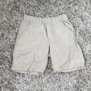 Beiga shorts från Lindex i linne - Snygga beiga shorts från Lindex i storlek 152. De har elastisk midja, två fickor fram och två bakfickor. Tillverkade i ett svalt linnematerial som passar perfekt till varma dagar. Enkla att matcha med det mesta och riktigt sköna att ha på sig.