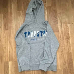 Grå hoodie från Trapstar med tryck - Säljer en grå hoodie från Trapstar med blått och vitt tryck på bröstet där det står 'TRAPSTAR'. Tröjan har huva med metallöljetter och klassisk känguruficka. Mjuk och skön bomullsblandning, perfekt för chill dagar.