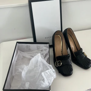 Svarta Gucci pumps med pärldetalj - Snygga svarta pumps från Gucci i skinn med chunky klack, dekorativ frans framtill och ikonisk GG-logo med pärlor i guld. Insidan har gulddetaljer och skon har en klassisk, lyxig vibe. Perfekta för dig som vill sticka ut med exklusiva detaljer. Använd ca 2 gånger 