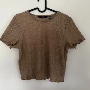 Beige ribbad topp från veromoda - Säljer en beige ribbad topp från veromodamed korta ärmar och vågiga kanter. Toppen har en croppad passform och är tillverkad i ett stretchigt material som sitter snyggt på kroppen. Perfekt att matcha med jeans eller kjol.