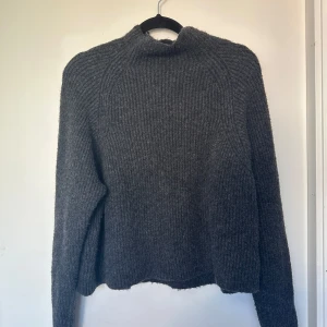Mörkgrå stickad tröja från Kappahl - Mysig mörkgrå stickad tröja från Kappahl med hög hals och ribbad struktur. Perfekt för kyliga dagar och enkel att matcha med jeans eller kjol. Långärmad och i en klassisk, lite loose passform.