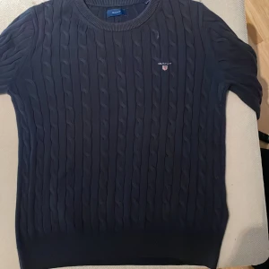 Mörkblå kabelstickad tröja från GANT - Snygg mörkblå kabelstickad tröja från GANT med rund halsringning och diskret logga på bröstet. Tröjan har korta ärmar och är tillverkad i mjuk bomull, perfekt för en stilren och klassisk look.