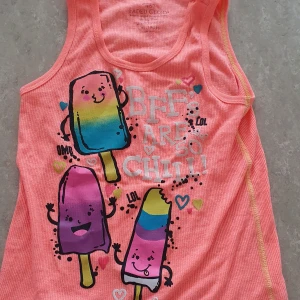 Linne - Säljer ett färgglatt rosa/korall linne med tryck av glada glassar i blått, gult, lila och rosa. Texten 'BFFs are so chill!' och små hjärtan gör linnet extra lekfullt. Perfekt för varma dagar och somrig stil.