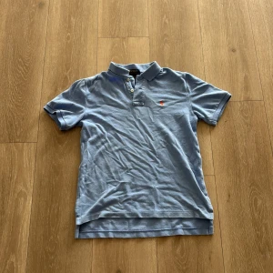 Blå Polo Ralph Lauren t-shirt - Snygg ljusblå t-shirt från Polo Ralph Lauren med klassisk krage och korta ärmar. T-shirten har knappar vid halsen och den ikoniska orangea broderade loggan på bröstet. Tillverkad i 100% bomull för skön känsla och stilren look.