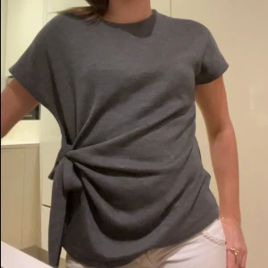 Mörkgrå topp med knyt i sidan - Säljer en mörkgrå topp med korta ärmar och snygg knytdetalj i sidan. Toppen har rund halsringning och är i ett mjukt, stickat material som känns skönt mot huden. Perfekt för dig som gillar stilrena plagg med en twist.
