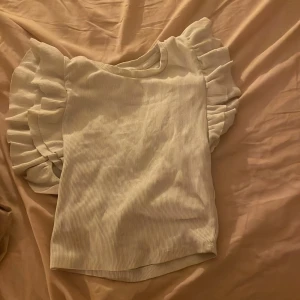 Vit ribbad topp med volangärmar - Supersöt vit ribbad topp med breda volangärmar som ger en extra touch till outfiten. Toppen är ärmlös och har en mjuk, stretchig känsla. Perfekt att styla med jeans eller kjol för en trendig look. Den har en smink fläck på ena hörnet men annars bra!