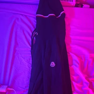 Svart vindjacka från Moncler med huva - Svart vindjacka från Moncler med klassisk logga på ärmen och snygga ränder i rött och vitt över bröstet. Jackan har huva med snörning och sportig vibe, perfekt för dig som gillar streetstyle. Materialet är lätt och känns bekvämt.