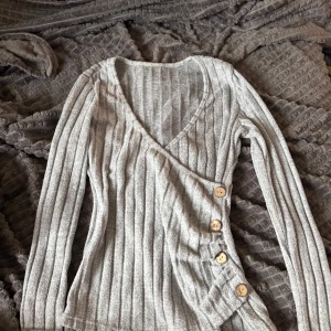 Grå ribbad omlottopp med knappar - Supersnygg grå ribbad omlottopp med v-ringning och dekorativa träknappar längs sidan. Långärmad och i mjukt material, perfekt för lager-på-lager-stil. Enkel att matcha med både jeans och kjol.