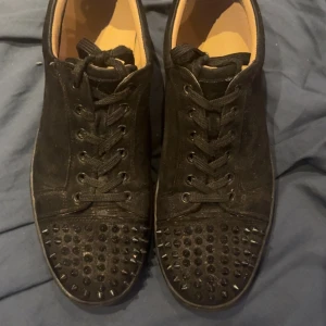 Christian louboutin svarta skor - Unika svarta sneakers i mocka från Christian Louboutin med coola svarta nitar på tån och den ikoniska röda sulan. Skorna har snörning och en klassisk rund tå. Perfekta för dig som vill sticka ut med en edgy vibe.