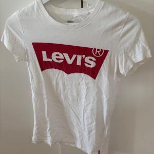 Vit Levi's t-shirt med röd logga - Vit t-shirt från Levi's med klassisk röd logga tryckt på bröstet. Rund hals och korta ärmar, tillverkad i mjuk bomull. Perfekt för dig som gillar enkel och ikonisk stil. 