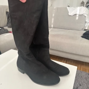 Svarta mockastövlar storlek 38 - Snygga svarta stövlar från Duffy i mocka-look med låg klack och rund tå. Klassisk design som går upp till knäet och dragkedja på insidan för enkel påtagning. Perfekta till höst- och vinteroutfits.