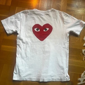 Vit t-shirt Comme des Garçons - Snygg vit t-shirt från Comme des Garçons PLAY med stort rött hjärta med ögon på både fram- och baksidan. Mindre hjärta med ögon på bröstet. Klassisk rund hals och korta ärmar. Tillverkad i mjuk bomull, perfekt för en chill och trendig look. Priset är  inte hugget i sten, frakt sker inom 12 timmar och hör av er om ni har frågor eller funderingar!
