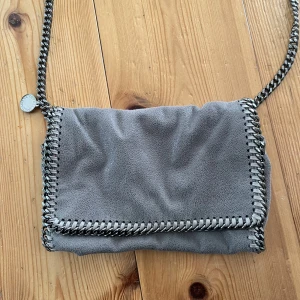 Grå väska från Stella McCartney - Säljer en snygg grå väska från Stella McCartney med kedjedetaljer längs kanterna och axelremmen. Väskan är väldigt trendig och kan bäras på flera olika sätt och passar till alla tillfällen. Jag tror att nypris ligger runt 5200. Kan även tänka mig byta. Pris kan diskuteras.