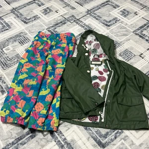 Grön regnjacka från Mango Kids - Snygg grön regnjacka från Mango Kids med huva och vita knappar. Insidan har ett coolt ananasmönster i rosa, gult och grönt. Jackan har dragkedja och stora fickor framtill. Perfekt för regniga dagar när du vill sticka ut lite extra.