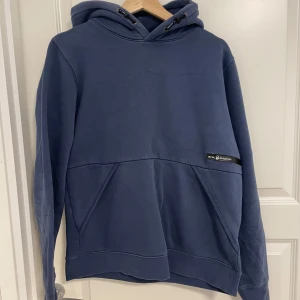 Blå hoodie från Sail Racing - Mörkblå hoodie från Sail Racing med huva och snörning. Tröjan har en stor magficka framtill och en liten logotyp på ena sidan. Tillverkad i mjuk bomullsmix, perfekt för kyliga dagar. Klassisk och stilren design som funkar till det mesta.