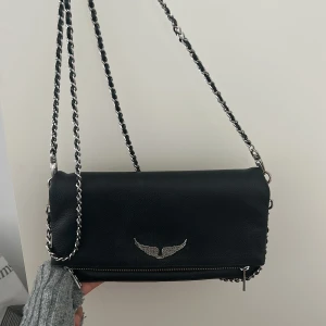 Svart Zadig & Voltaire axelväska i skinn - Cool svart axelväska från Zadig & Voltaire i skinn med silverfärgad kedjerem och snyggt vingemblem framtill. Väskan har dragkedja både fram och bak, perfekt för att hålla koll på dina grejer. Stilren och edgy design som lyfter vilken outfit som helst.