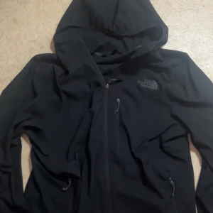 Svart vindjacka från The North Face - Svart vindjacka från The North Face med huva och dragkedja framtill. Jackan har flera praktiska fickor med dragkedja och en clean look. Perfekt för dig som vill ha en stilren och funktionell jacka till vardags. Klassisk logga på bröstet.