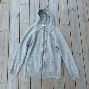 Grå kashmirhoodie från Davida - Supermjuk grå hoodie från Davida i 100% kashmir. Tröjan har dragkedja, huva med snörning och två fickor framtill. Perfekt för dig som gillar en chill och lyxig stil. Långärmad och i klassisk passform. Sitter som S.