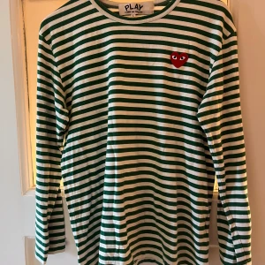 Randig långärmad tröja Comme des Garçons - Grön- och vitrandig långärmad tröja från Comme des Garçons PLAY med klassiskt rött hjärta på bröstet. Mjuk bomull, rund hals och relaxed fit. Perfekt för dig som gillar streetstyle och vill sticka ut med en ikonisk detalj.