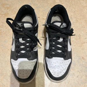 Nike Dunk Low svart/vit sneakers - Nike Dunk Low sneakers i svart och vitt med klassisk Swoosh på sidan. Skorna har snörning, perforerad tå och låg profil. Ovandelen är i mocka och syntet, med svart foder och vit sula. Perfekta för dig som gillar streetstyle och vill ha en ikonisk look.