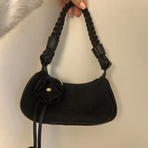 Svart handväska - Jätt jättenygg svart vintage axelväska med flätat handtag och superfin blomdetalj framtill. Liten och perfekt att ta med på t.ex konserter med väskförbud, men stor nog att få plats med min stora mobil plus mycket mer! Den har såå fina detaljer💕