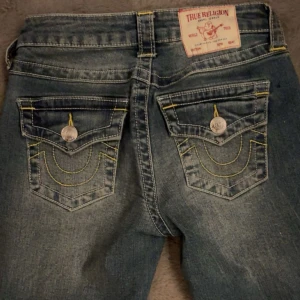 True Religion Becca Bootcut Jeans - Säljer ett par True Religion Becca jeans i klassisk blå tvätt med coola kontrastsömmar i gult. Modellen är midwaist och bootcut, med snygga fickdetaljer och silverfärgade knappar. Jeansen är i stretchig bomull och har en riktigt schysst passform. Knappast använda och i nyskick!!