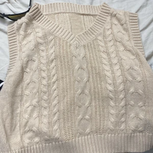 Beige stickad kabelväst med v-ringning - Säljer en beige stickad väst med klassiskt kabelstickat mönster och v-ringad hals. Västen har breda ribbade kanter vid hals och ärmhål, vilket ger en snygg och chill look. Perfekt att slänga över en skjorta eller t-shirt för extra stilpoäng.